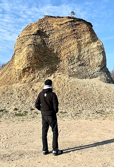 A young man dressed in all black - jeans, jacket and Addidas shoes - looks at a clay mountain over a blue sky.